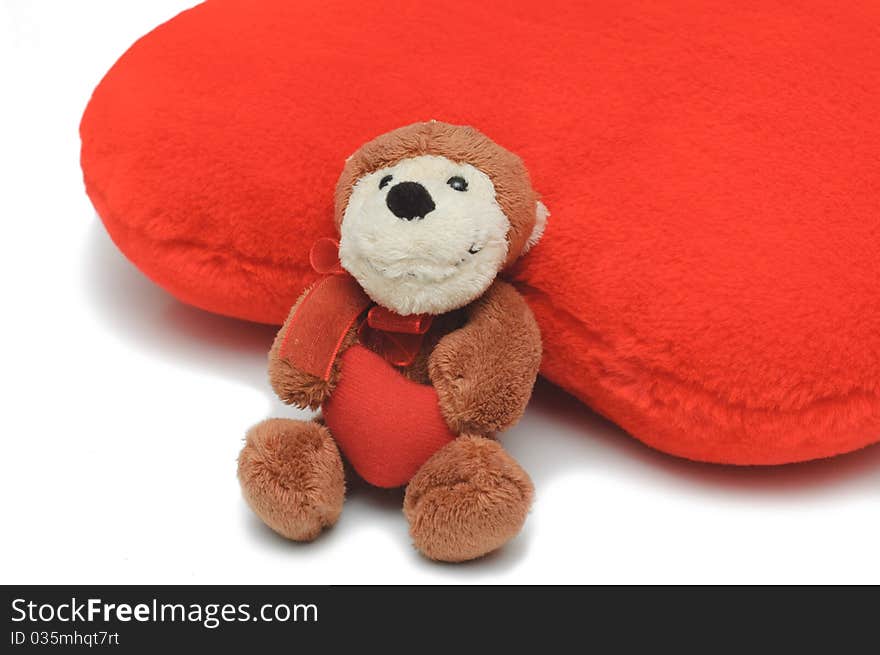 Little teddy-bear holding red heart. Bigger red pillow-heart behind it. White isolated background