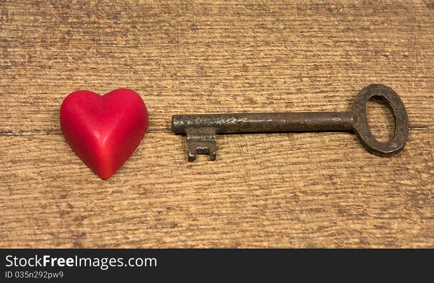 On the image there is old key and little red heart on the old wooden background.