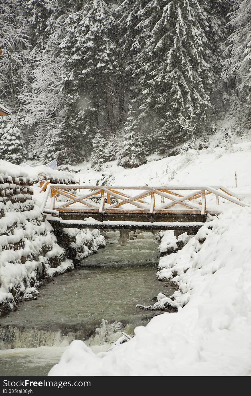 The bridge through the mountain river near wood, under going snow. The bridge through the mountain river near wood, under going snow