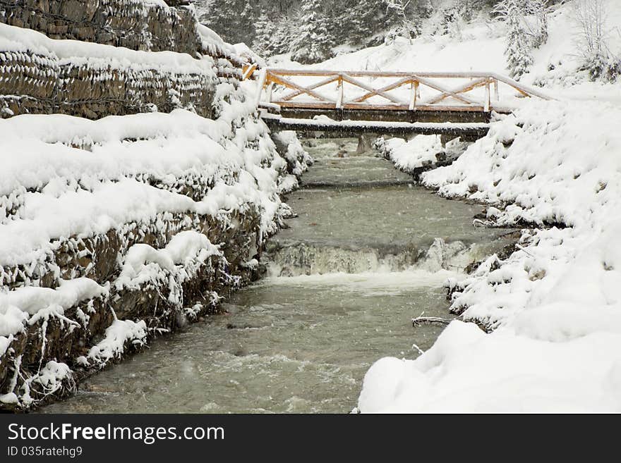 The bridge through the mountain river near wood, under going snow. The bridge through the mountain river near wood, under going snow