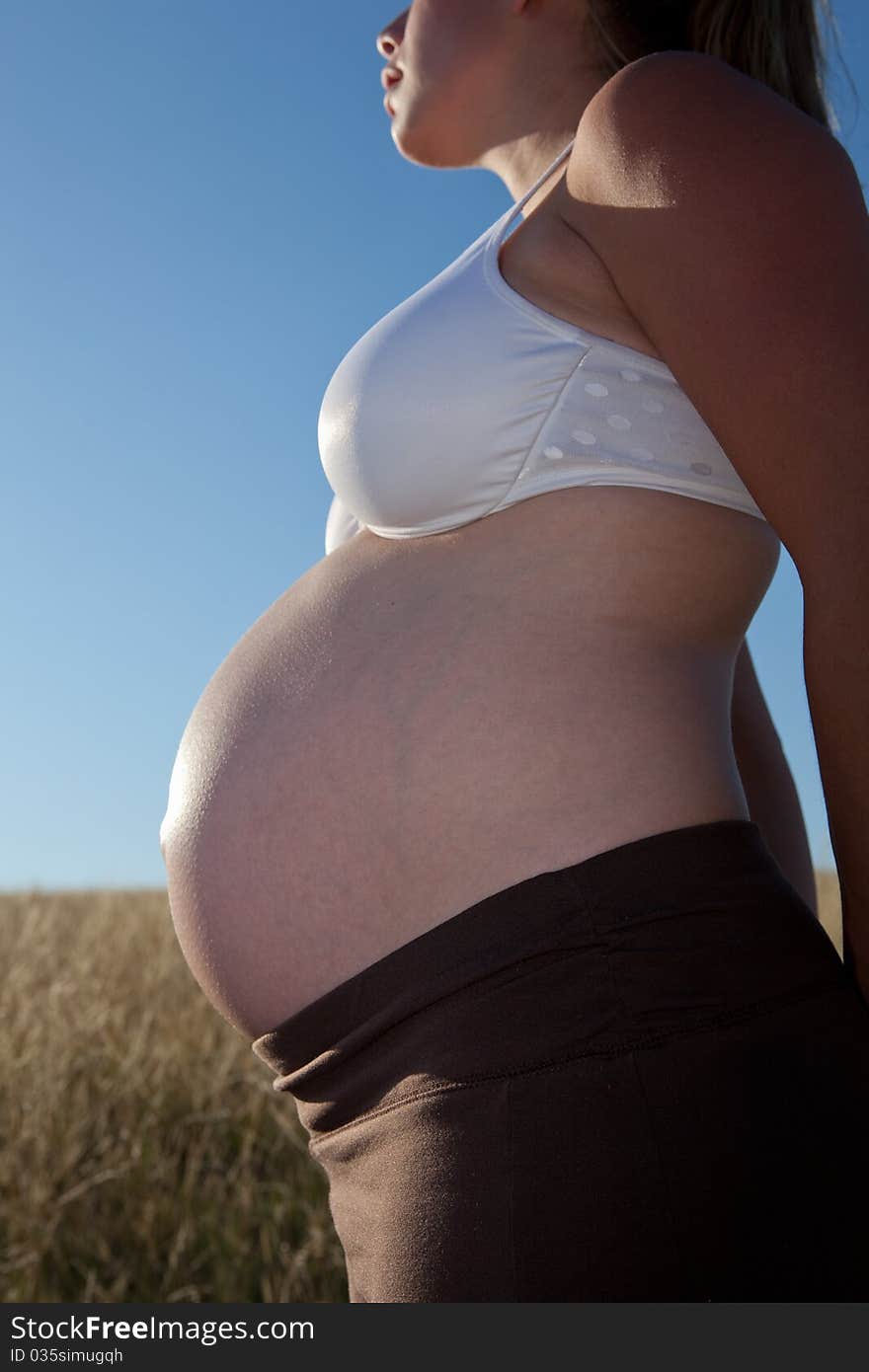 A young pregnant woman standing in an open field wearing a bra and looking peaceful.