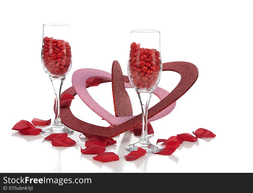 A front closeup view of two champagne glasses filled with cinnamon hearts, with multi-sized glitter valentines and rose petals on the white background. A front closeup view of two champagne glasses filled with cinnamon hearts, with multi-sized glitter valentines and rose petals on the white background.