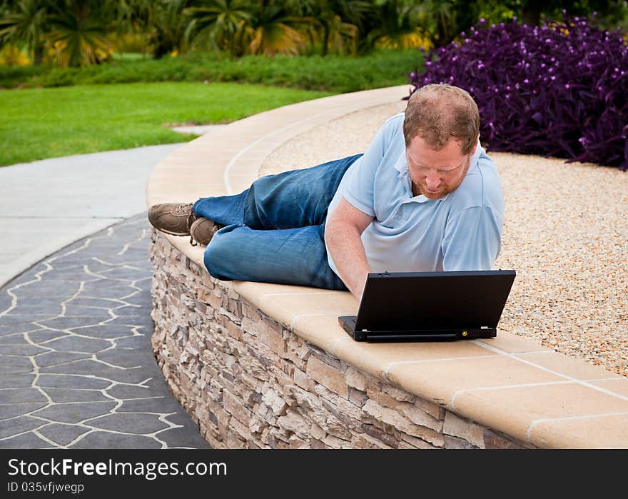 Man lying down on a wall in a park using a laptop computer. Man lying down on a wall in a park using a laptop computer.