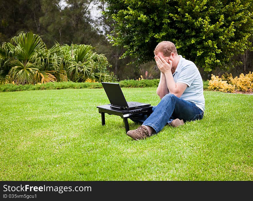 Man displaying emotion with his hands to his face in front of a laptop computer in the park. Man displaying emotion with his hands to his face in front of a laptop computer in the park.