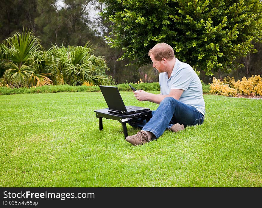 Man sitting in a park with a laptop and sending an SMS on his cell phone. Man sitting in a park with a laptop and sending an SMS on his cell phone.