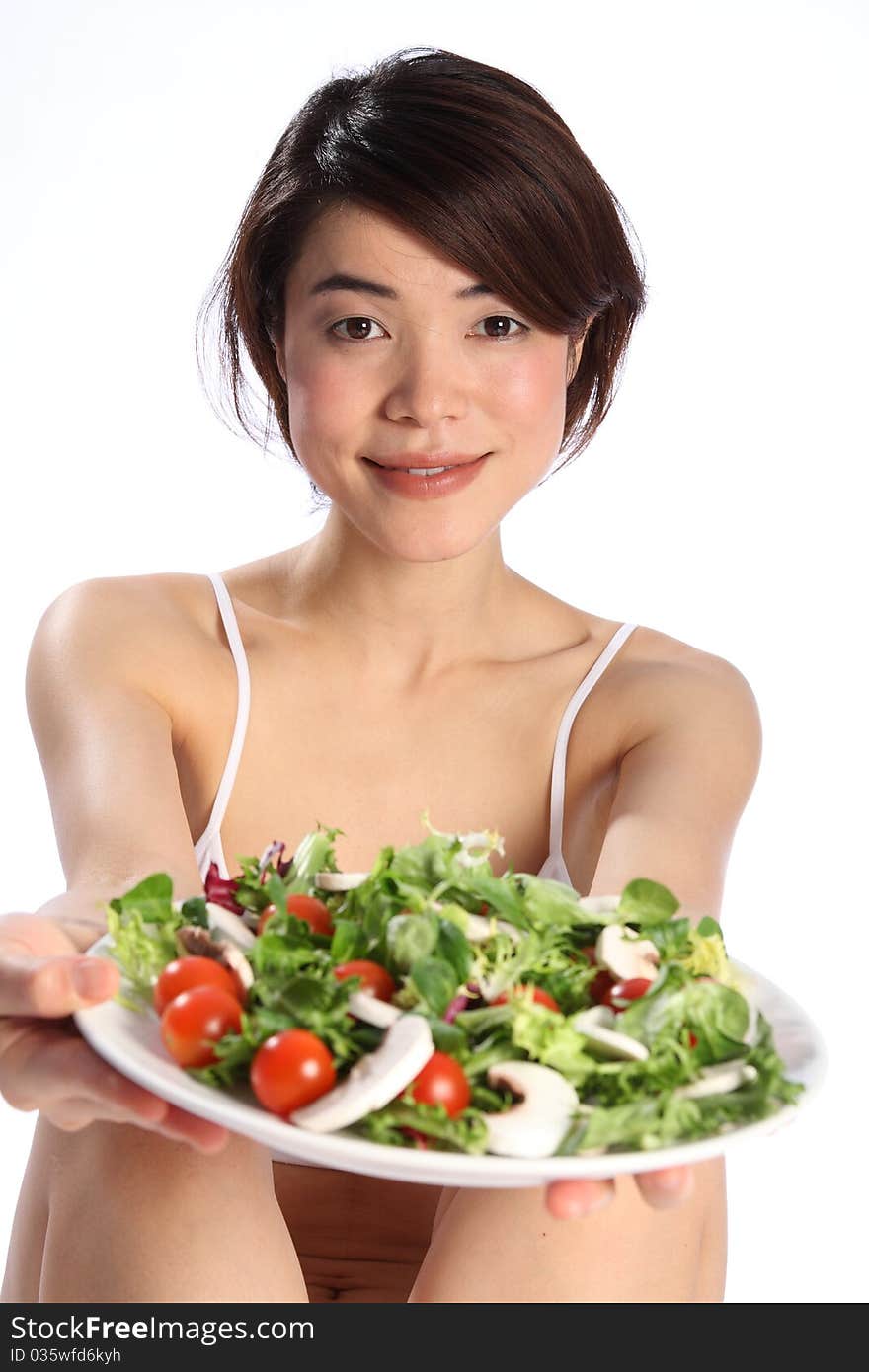 Beautiful, smiling young Japanese girl, wearing white sports bra, holds out a plate of green salad. Focus is sharp on model with salad out of focus in foreground. Beautiful, smiling young Japanese girl, wearing white sports bra, holds out a plate of green salad. Focus is sharp on model with salad out of focus in foreground.