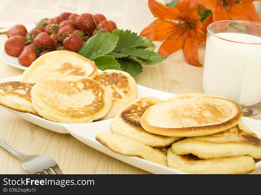 On the table a plate of pancakes, a plate of strawberries, a glass of milk and orange lilies. Breakfast