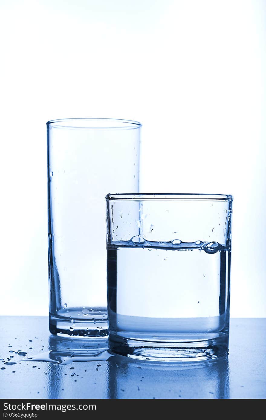 Empty glass behind full glass of water. Image stylized in blue tone for more cool feeling. Empty glass behind full glass of water. Image stylized in blue tone for more cool feeling