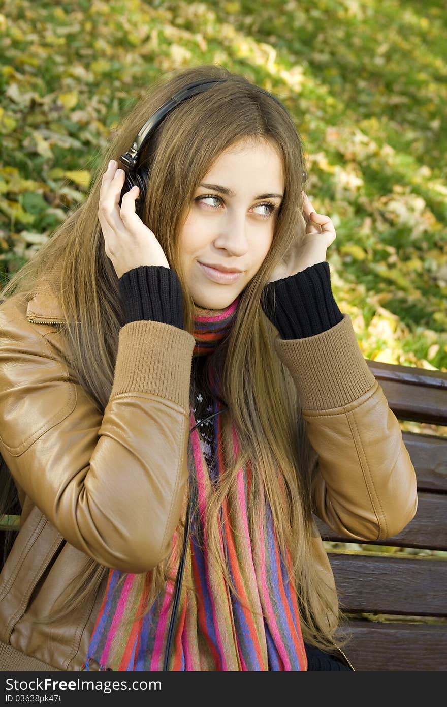 Young Caucasian woman with headphones in autumn park sitting on a wooden bench. Autumn around a lot of colorful foliage. Young Caucasian woman with headphones in autumn park sitting on a wooden bench. Autumn around a lot of colorful foliage