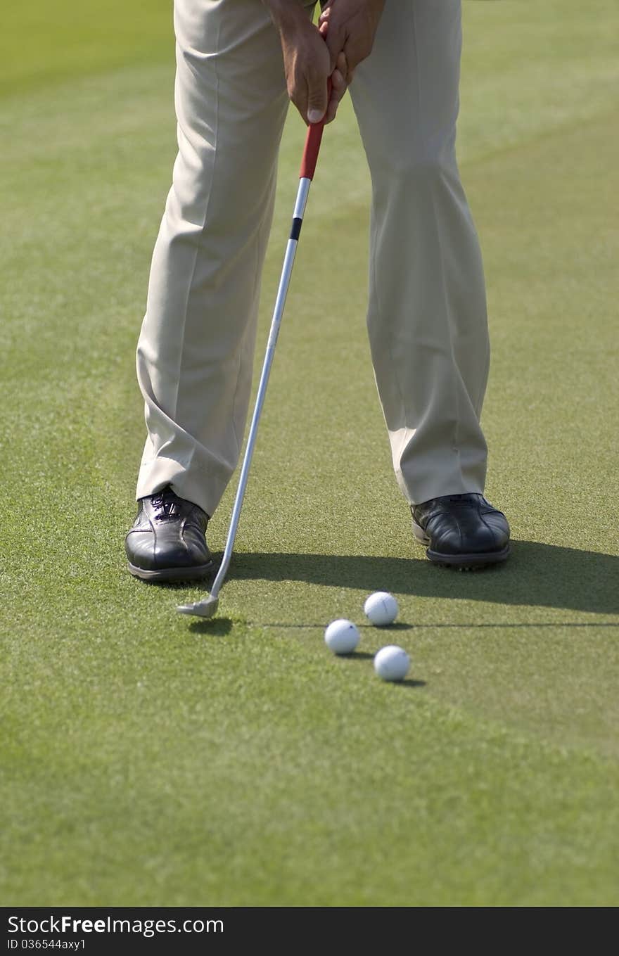 A golfer wearing beige pants and black leather shoes practicing his putt with several balls on a sunny day. A golfer wearing beige pants and black leather shoes practicing his putt with several balls on a sunny day