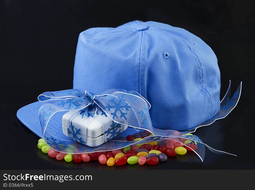 Blue baseball cap, blue ribbon around a white jewelry box placed on colorful jellybeans on black background reflect the sweet romance of love and sports. Blue baseball cap, blue ribbon around a white jewelry box placed on colorful jellybeans on black background reflect the sweet romance of love and sports