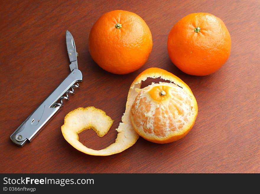 Three Mandarin Oranges, one of which is peeled, on a dark wood background with a pen knife