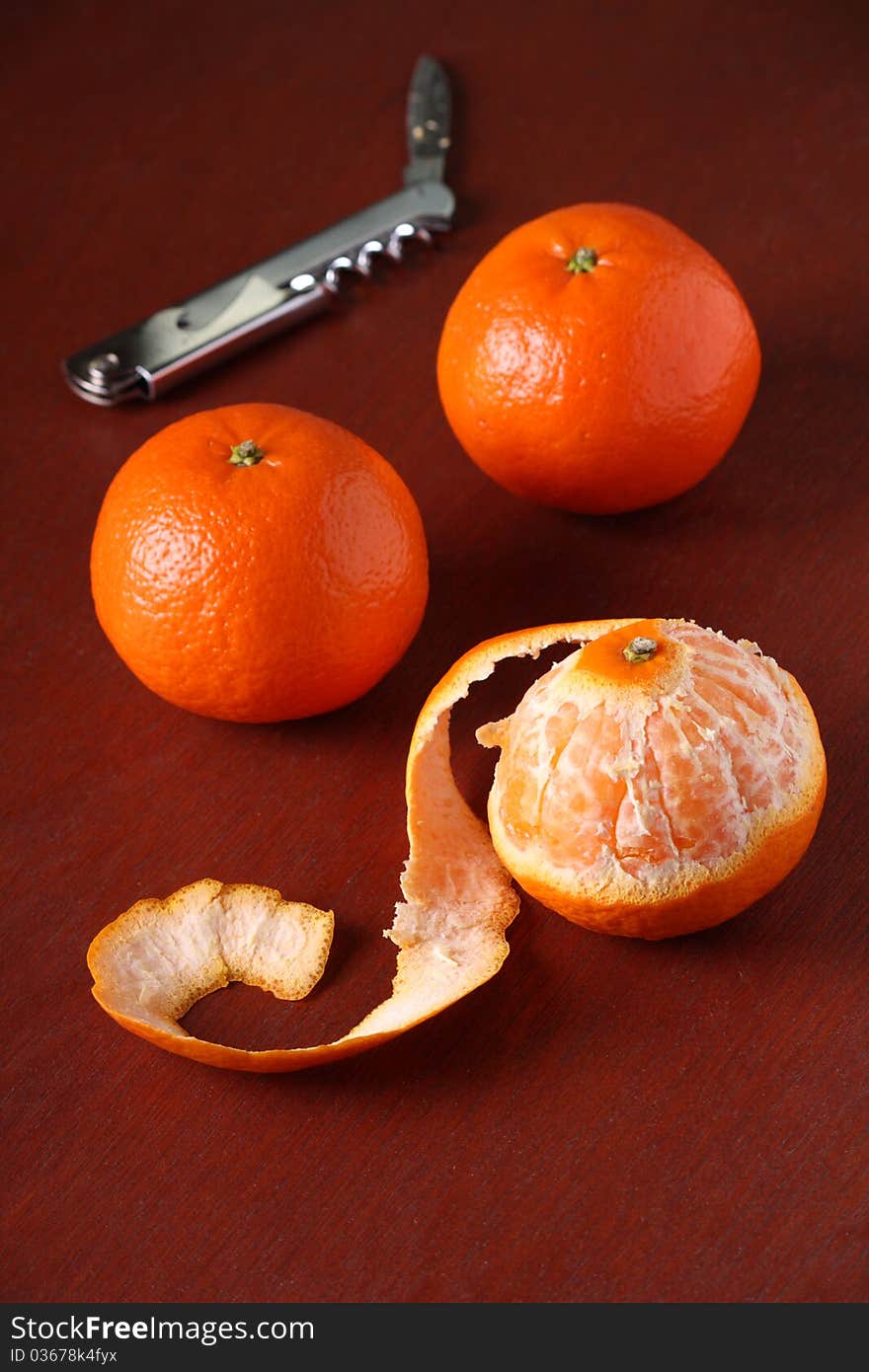 Three Mandarin Oranges, one of which is peeled, on a dark wood background with a pen knife