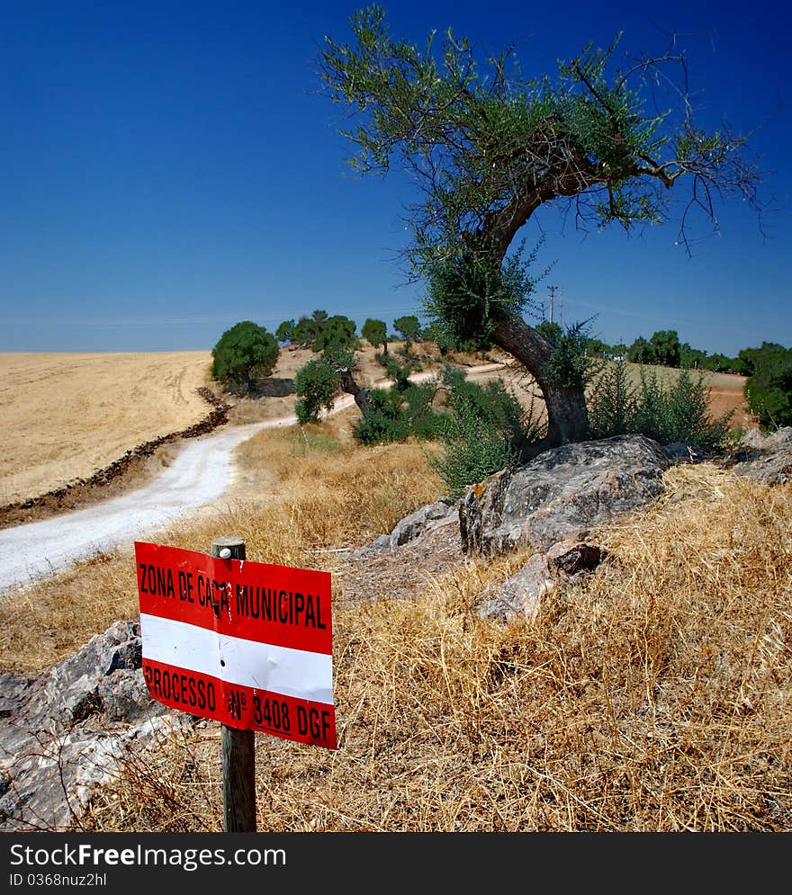 White and red sign with a country road under blue sky. Trees on fields with grass burned by the sun. White and red sign with a country road under blue sky. Trees on fields with grass burned by the sun