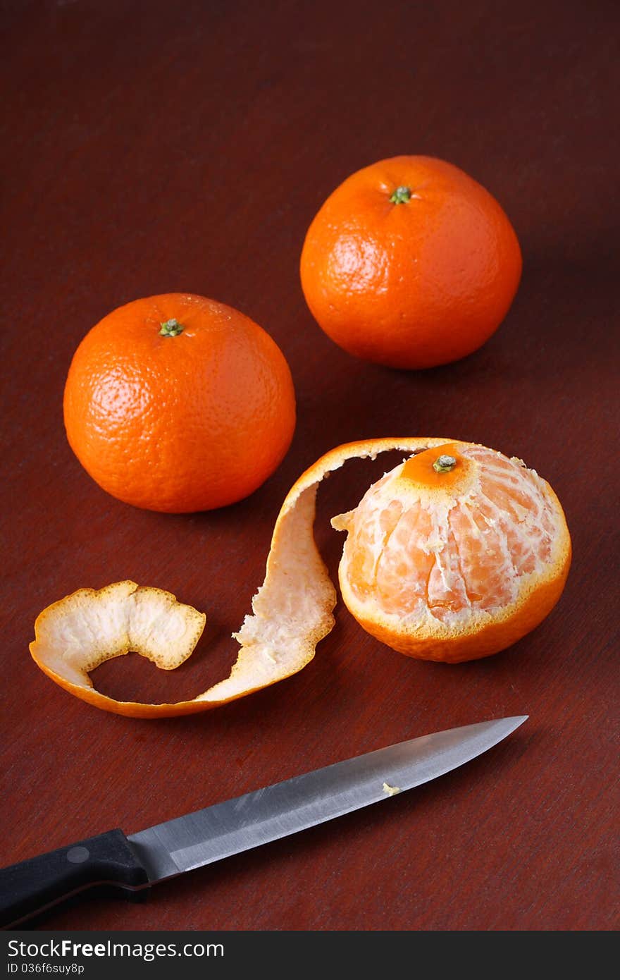 Three Mandarin Oranges, one of which is peeled, on a dark wood background with a kitchen knife