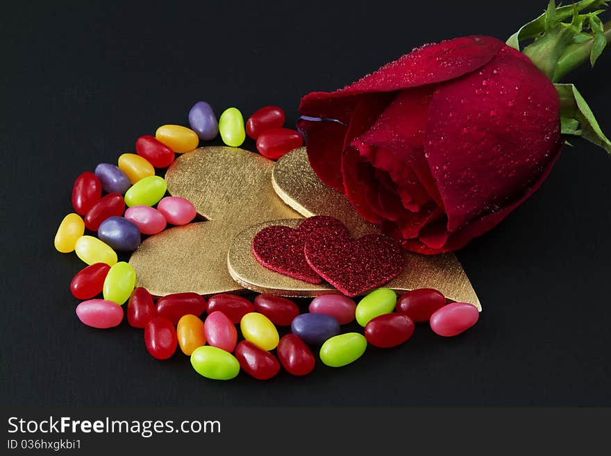 A single, red rose is placed on a black background with wo gold hears, two red hearts, and colorful jelly beans;. A single, red rose is placed on a black background with wo gold hears, two red hearts, and colorful jelly beans;