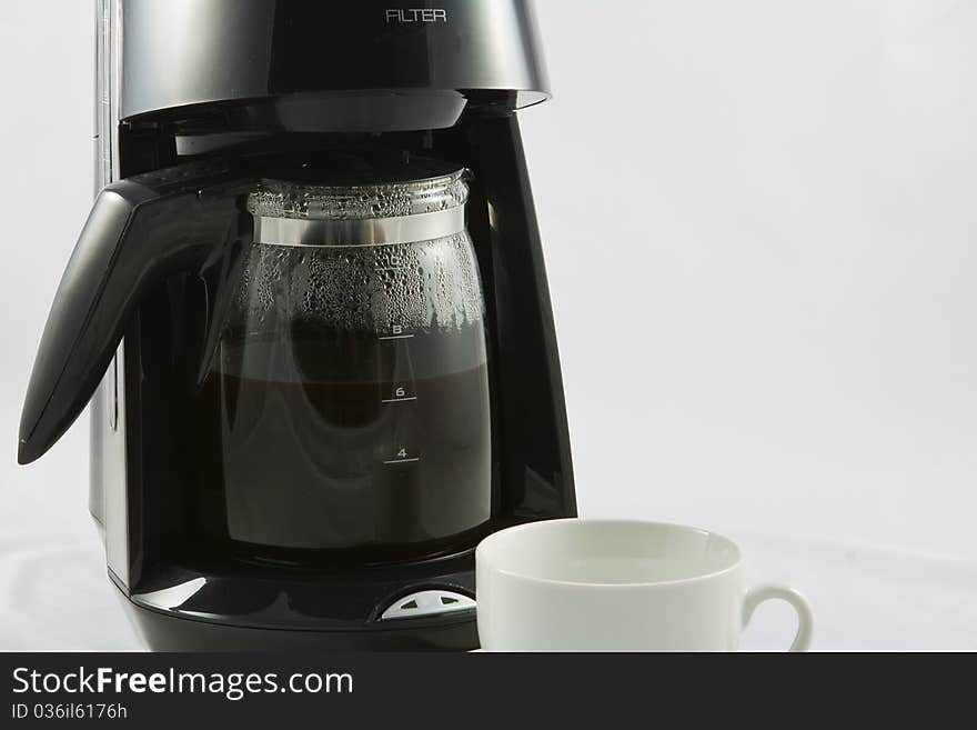 A black coffee machine with a glass jug full of hot freshly made coffee and a white cup waiting to be filled up. A black coffee machine with a glass jug full of hot freshly made coffee and a white cup waiting to be filled up