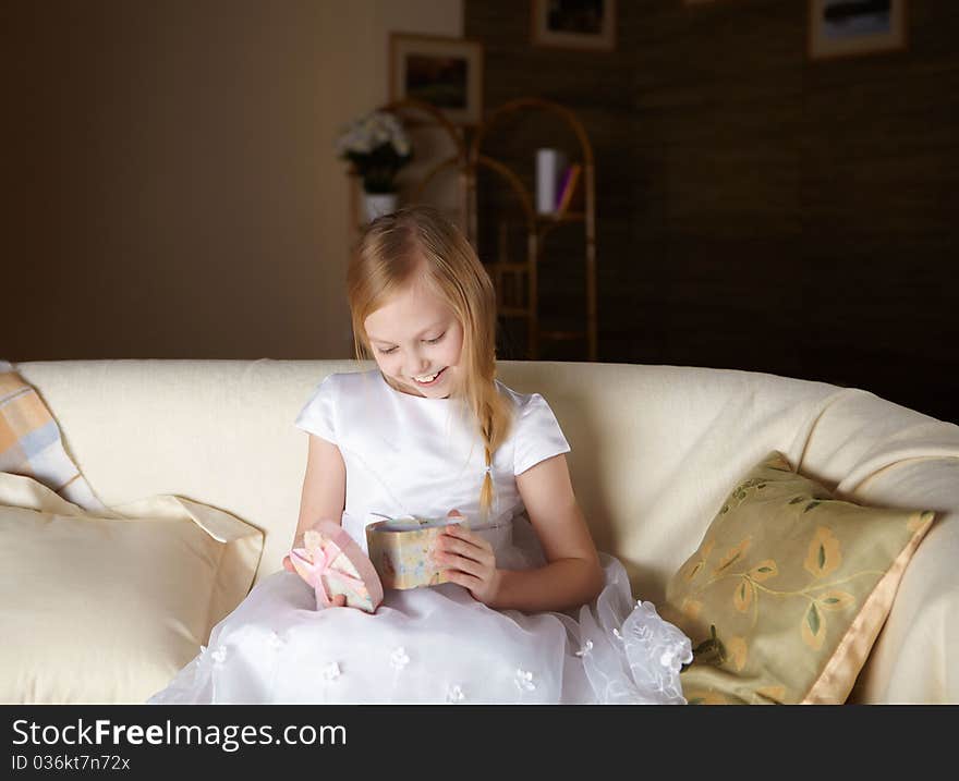 A beautiful little girl in white dress sitting on a sofa with a present at home. A beautiful little girl in white dress sitting on a sofa with a present at home