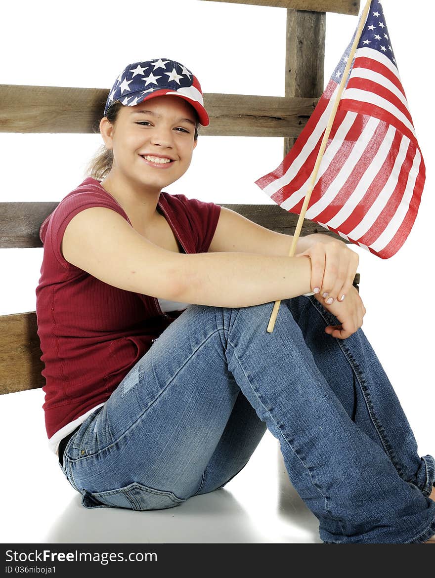 A pretty young teen sitting against a fence, wearing a stars-and-stripes hat and holding an American flag. Isolated on white. A pretty young teen sitting against a fence, wearing a stars-and-stripes hat and holding an American flag. Isolated on white.