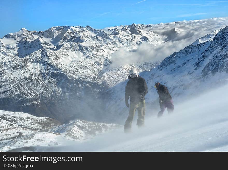 Two snowboarders on steep slope in very windy conditions. Scenic view of wonderful Austrian alps. Mountains covered in clouds in the background. Two snowboarders on steep slope in very windy conditions. Scenic view of wonderful Austrian alps. Mountains covered in clouds in the background.