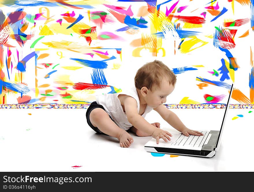 Little baby boy is sitting on floor with his laptop, over white wall, in messy painted room with many colors around