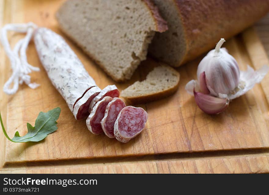 Dry sausage on a wooden cutting board with slices cut off and a knife. A bread is present in background. Dry sausage on a wooden cutting board with slices cut off and a knife. A bread is present in background.