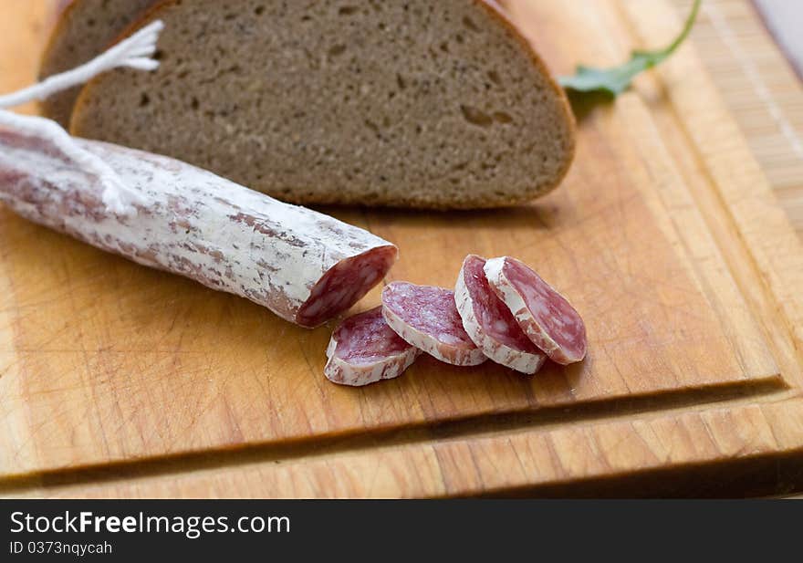 Dry sausage on a wooden cutting board with slices cut off and a knife. A bread is present in background. Dry sausage on a wooden cutting board with slices cut off and a knife. A bread is present in background.