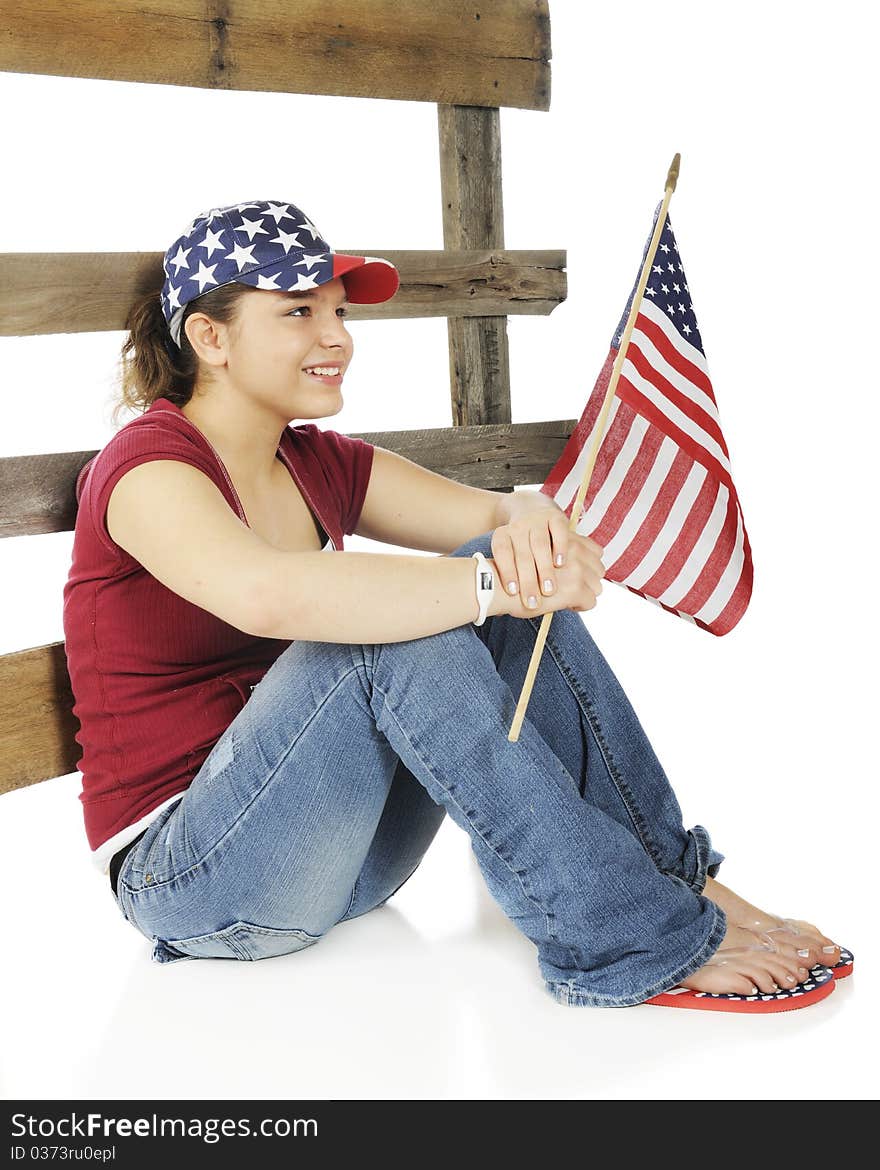 An attractive young teen holding an American flag and wearing a stars and stripes hat sitting against a rustic rail fence. Isolated on white. An attractive young teen holding an American flag and wearing a stars and stripes hat sitting against a rustic rail fence. Isolated on white.