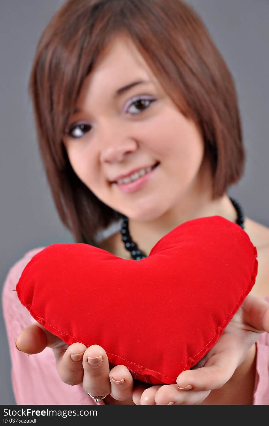 The beautiful young woman holds in hands a red heart on a grey background. Selective focus on heart.