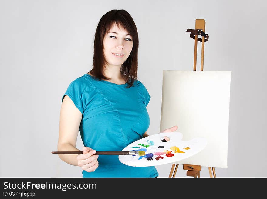 Young girl in blue shirt standing near easel, holding brush and palette, smiling. Young girl in blue shirt standing near easel, holding brush and palette, smiling