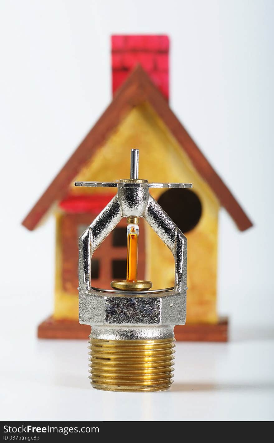 Close up image of fire sprinkler on white. Fire sprinklers are part of an integrated water piping system designed for life and fire safety. Replica of house in background. Close up image of fire sprinkler on white. Fire sprinklers are part of an integrated water piping system designed for life and fire safety. Replica of house in background