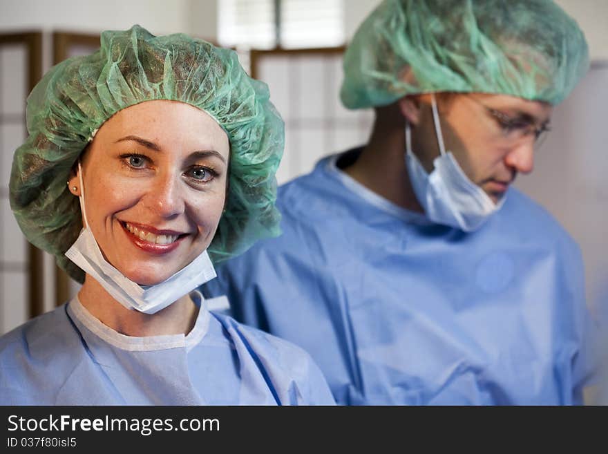 A lovely nurse with her surgical mask away from her face smiles at the camera with an out of focus doctor in the background. A lovely nurse with her surgical mask away from her face smiles at the camera with an out of focus doctor in the background.