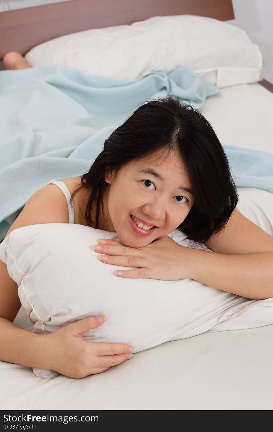 A smiling Asian woman in bed hugging a pillow while looking at you