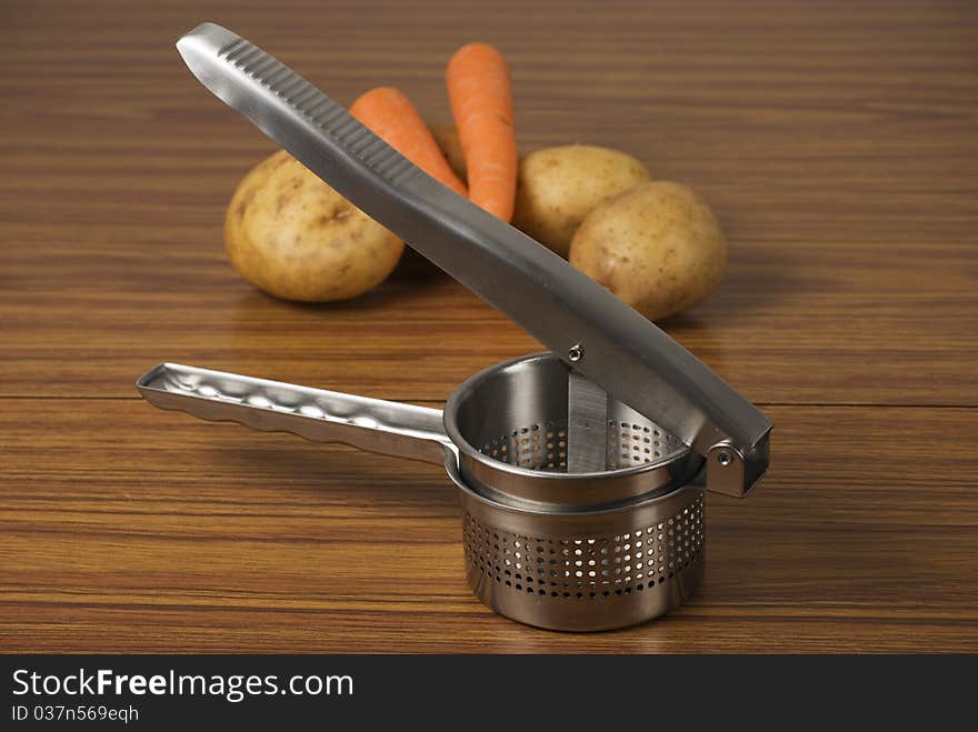 A horizontal image of a food ricer masher and vegetables on a wooden table. A horizontal image of a food ricer masher and vegetables on a wooden table