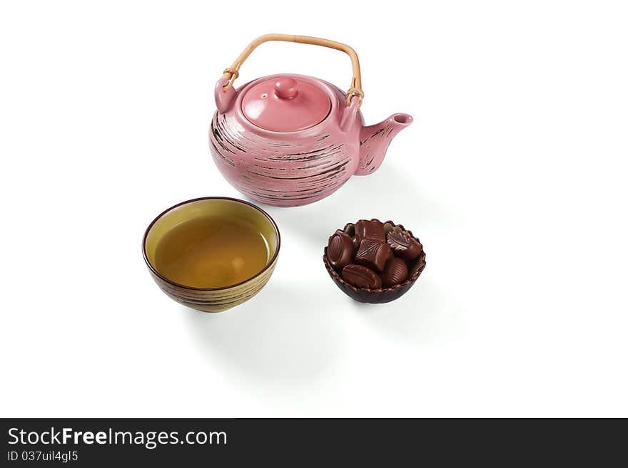 Composition with tea set that shows teakettle, tea in a cup and candies in a bowl on a white background with shadows.