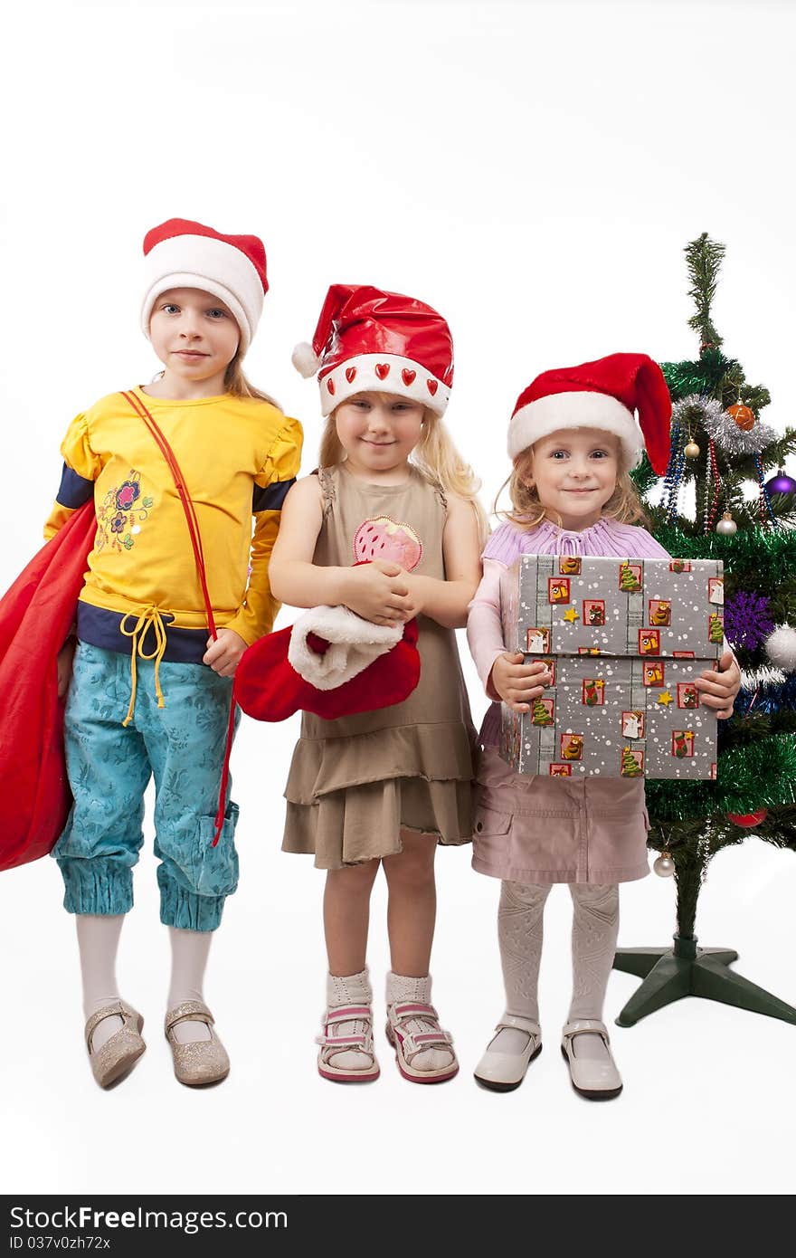 Three young little caucasian blond girls standing together near christmas tree holding gifts and smiling sincerely.isolated over white background. Three young little caucasian blond girls standing together near christmas tree holding gifts and smiling sincerely.isolated over white background