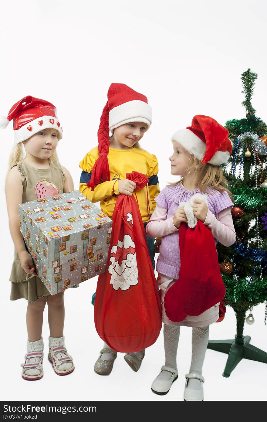 Three young little caucasian blond girls standing together near christmas tree holding gifts and smiling sincerely.isolated over white background. Three young little caucasian blond girls standing together near christmas tree holding gifts and smiling sincerely.isolated over white background