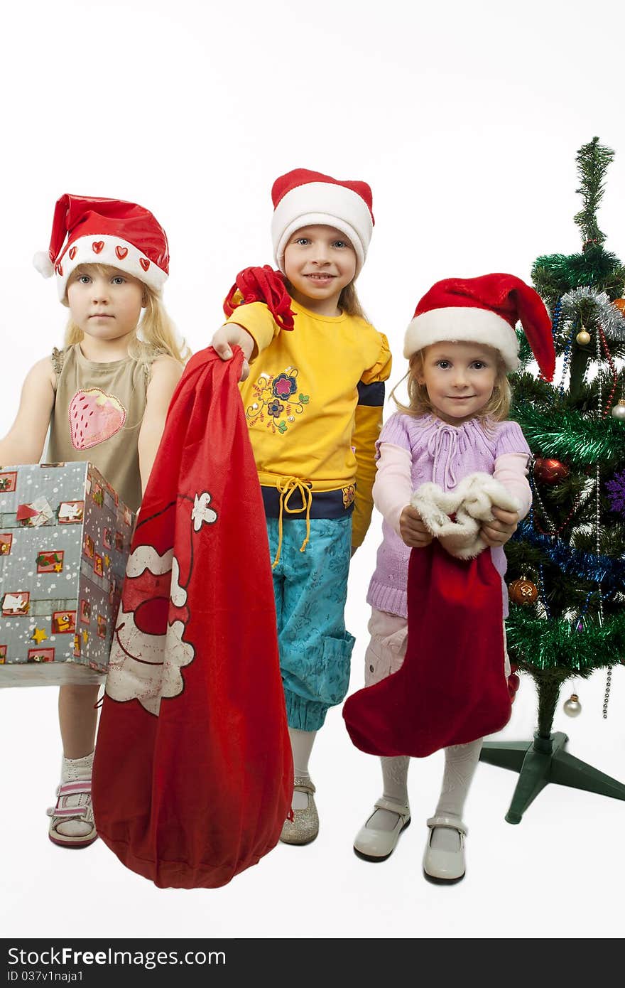 Three young little caucasian blond girls standing together near christmas tree holding gifts and smiling sincerely.isolated over white background. Three young little caucasian blond girls standing together near christmas tree holding gifts and smiling sincerely.isolated over white background