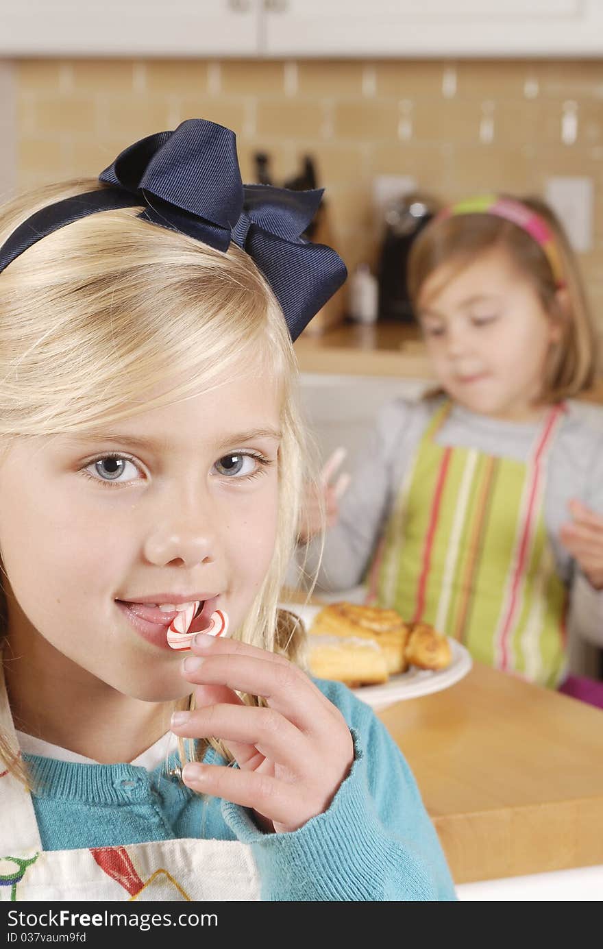 Sisters cooking cookies and cinnamon buns. Young girl with candy cane in foreground and sister in background holding a candy cane. Sisters cooking cookies and cinnamon buns. Young girl with candy cane in foreground and sister in background holding a candy cane.