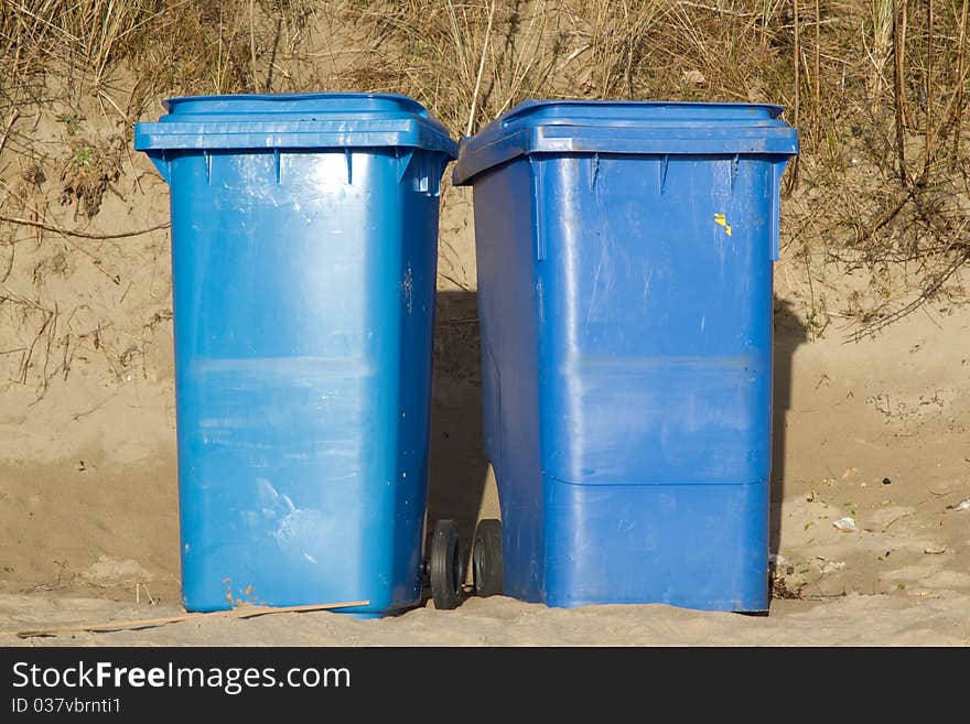 Two blue wheelie bins standing side by side on sand with grasses in the background. Two blue wheelie bins standing side by side on sand with grasses in the background.