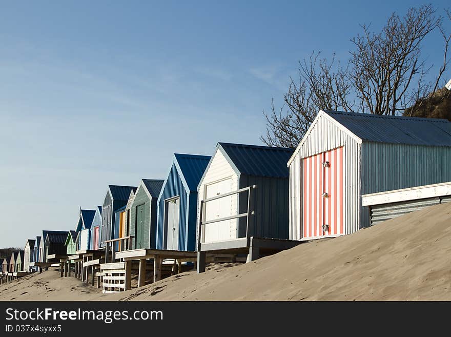 Beach huts, multicoloured on a sand dune, extend into the distance with a blue sky. Beach huts, multicoloured on a sand dune, extend into the distance with a blue sky.