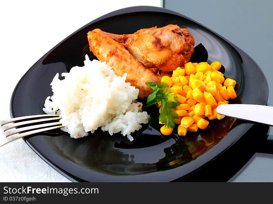 A piece of roast chicken with rice and corn on the black plate. A piece of roast chicken with rice and corn on the black plate