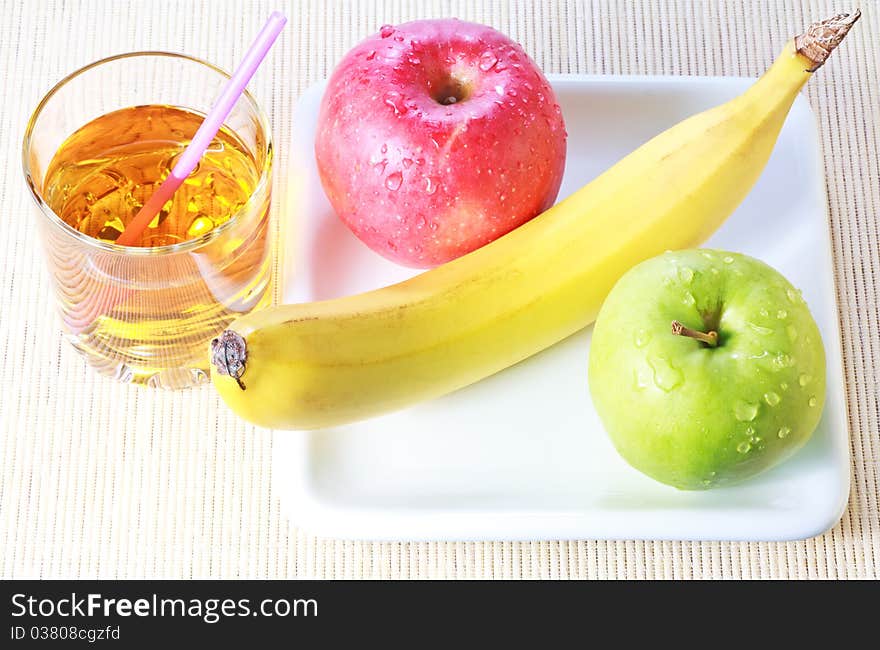 Banana, green and red apples on white plate and glass of apple juice over bamboo mat background. Banana, green and red apples on white plate and glass of apple juice over bamboo mat background