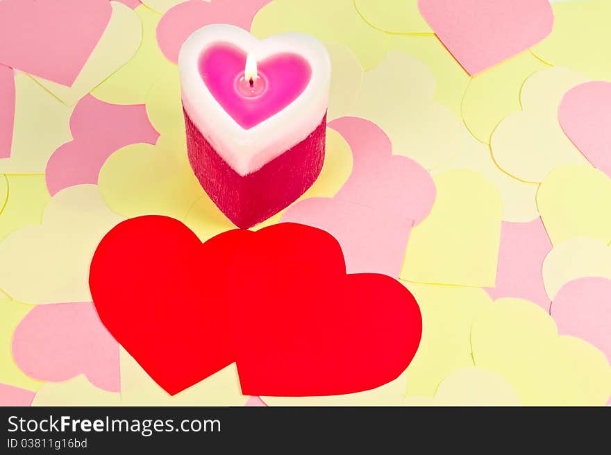 A heart shaped lighted candle over cute paper hearts background and two red hearts in the foreground. A heart shaped lighted candle over cute paper hearts background and two red hearts in the foreground.