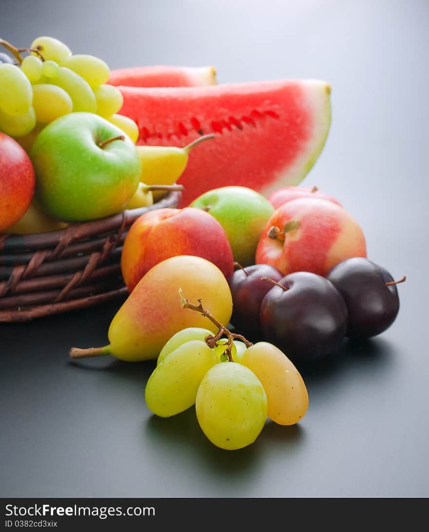 Various fresh ripe fruits placed in a wicker basket and around on gray gradient background