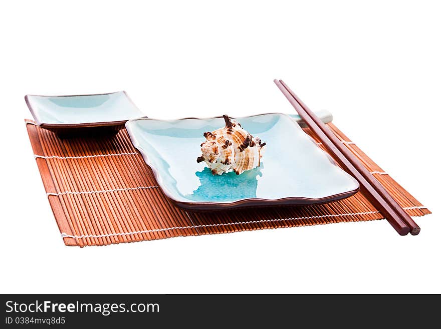 Traditional Japanese sushi tableware set: chopsticks on a rest, plate with a sea shell on it and a saucer, all on a bamboo place mat, on a white background, isolated. Traditional Japanese sushi tableware set: chopsticks on a rest, plate with a sea shell on it and a saucer, all on a bamboo place mat, on a white background, isolated.