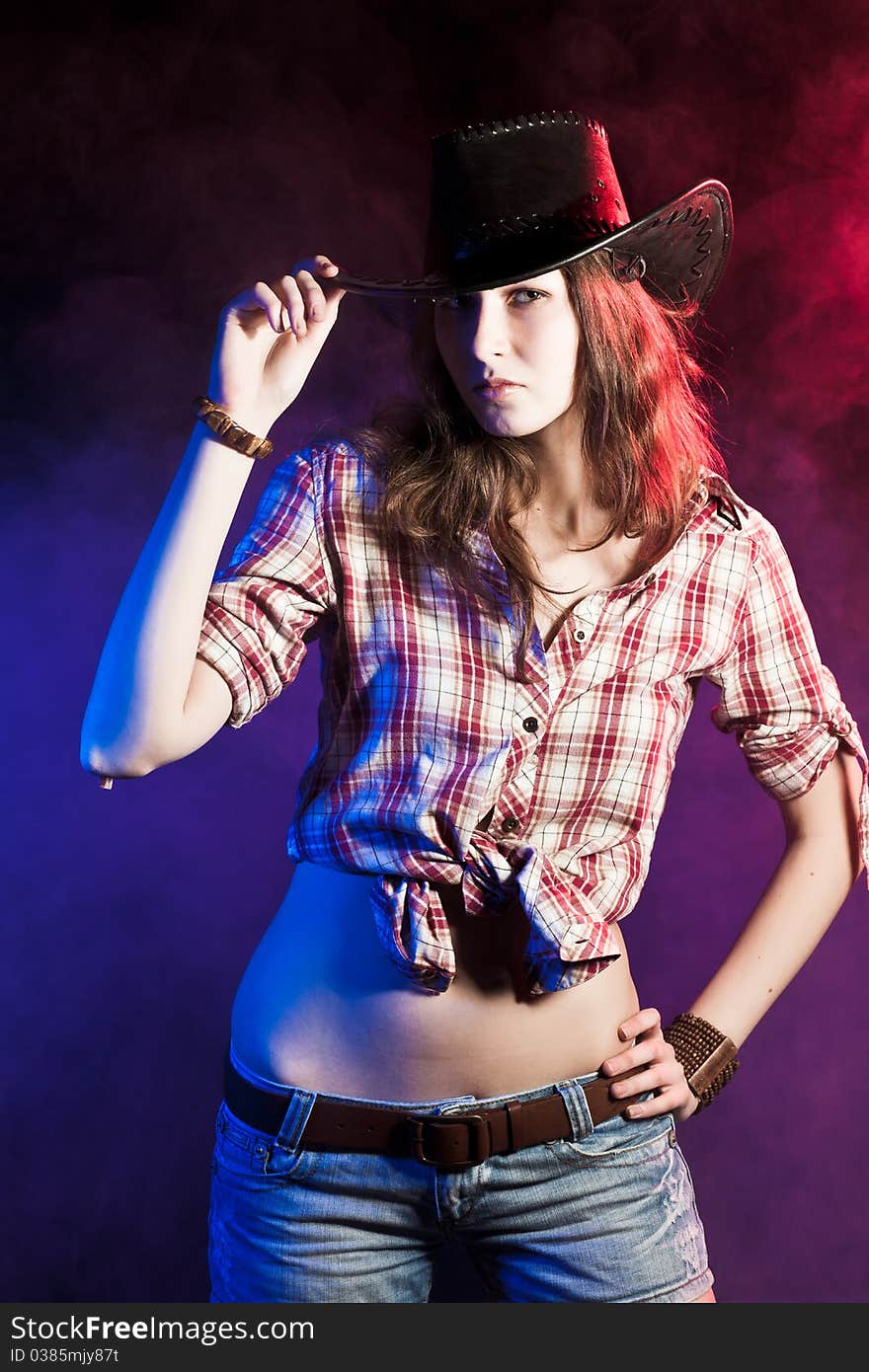 Studio portrait of a young pretty cowgirl in a checkered shirt, jeans shorts and black leather hat on a color smoky background. Studio portrait of a young pretty cowgirl in a checkered shirt, jeans shorts and black leather hat on a color smoky background