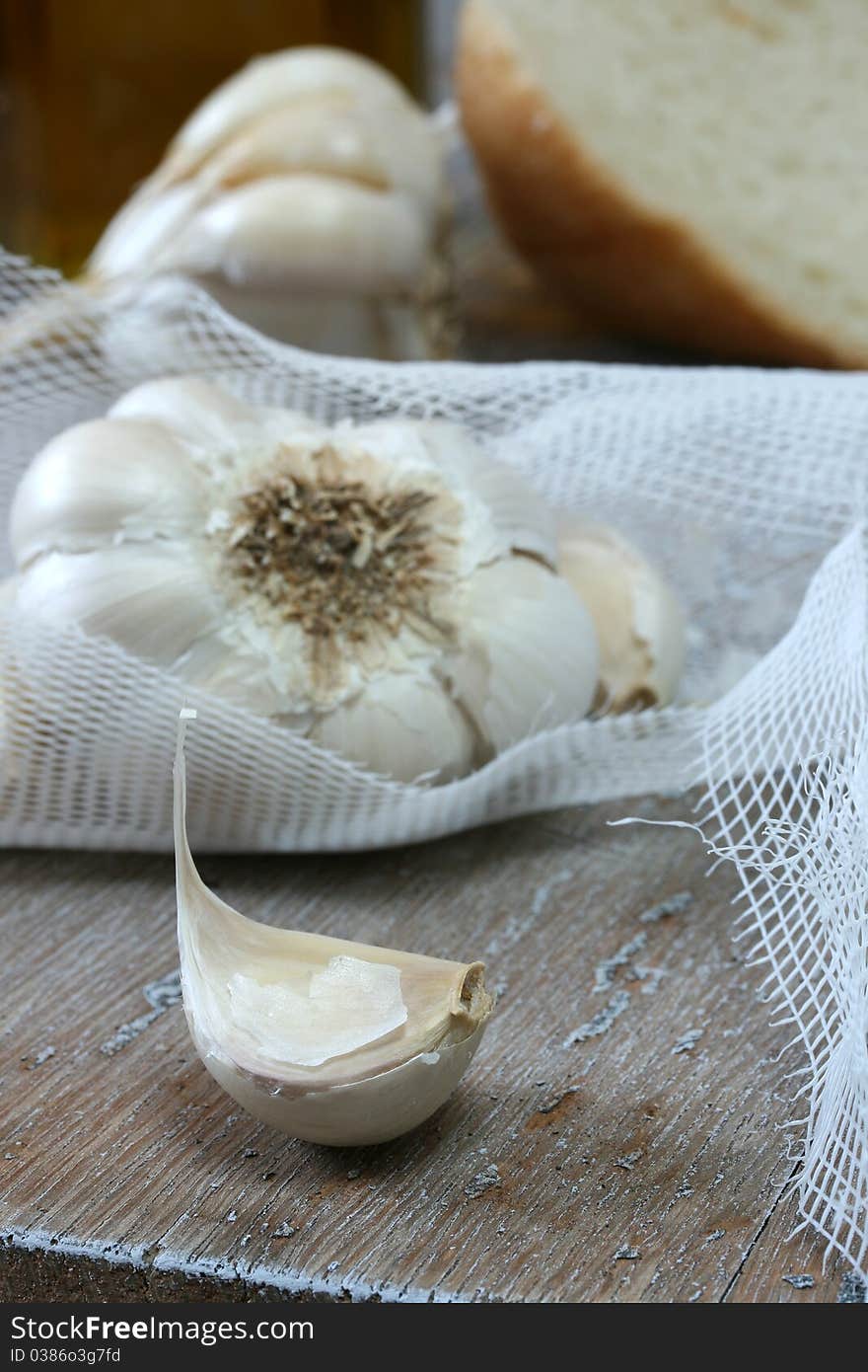 Clove and head of garlic in netting; more garlic, olive oil and bread in background. Clove and head of garlic in netting; more garlic, olive oil and bread in background