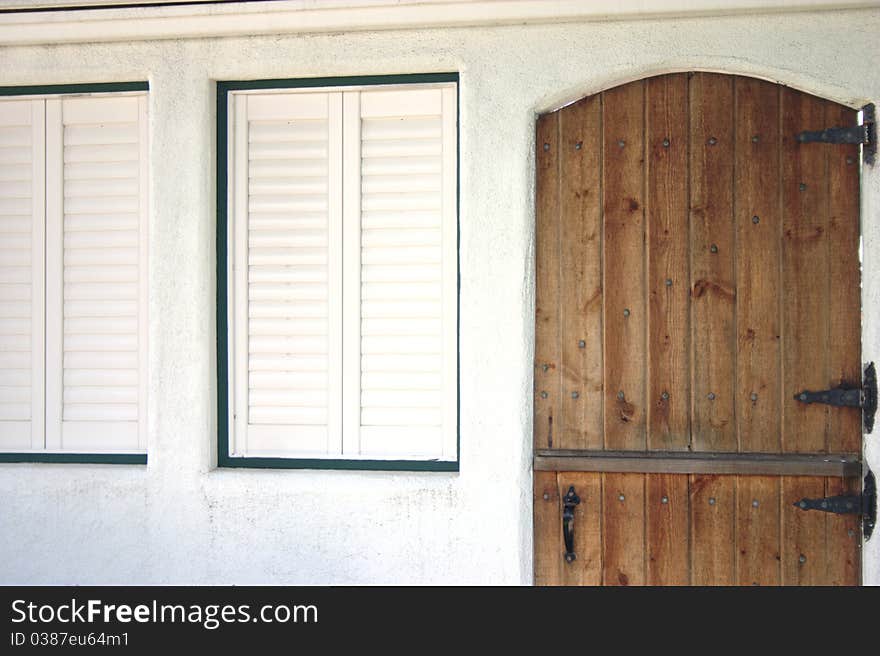 Stained wooden door with black hinges next to white painted shutters and green trim in a stucco wall. Stained wooden door with black hinges next to white painted shutters and green trim in a stucco wall