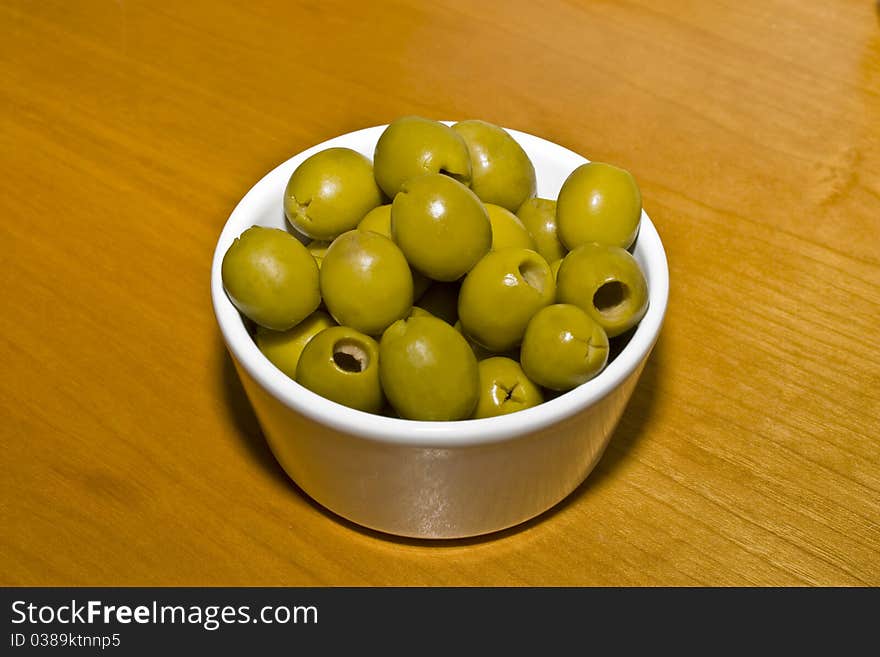 A small white china bowl on a wooden table containing green olives